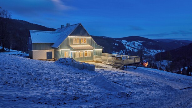 Une maison moderne dotée d’un grand porche et d’un espace abrité, entourée de collines enneigées et avec une chaîne de montagnes en arrière-plan.