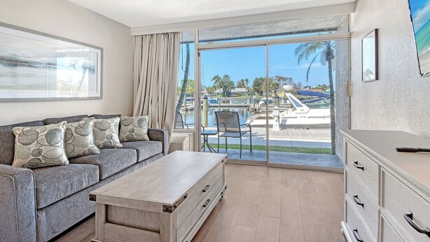 A modern living room with a grey sofa, a wooden coffee table, and a view of a marina through large glass doors. - Waterfront Hotel Room, Huge Pool, Tiki Bar, Close to Beaches & Great Food! (Sarasota)