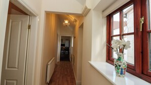 A narrow hallway with wooden flooring, a radiator, and a window with a vase of white flowers. - Partridge Cottage (Filey)