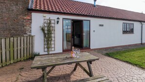 A white building with a red-tiled roof, a wooden fence, and a picnic table with wine glasses. - Partridge Cottage (Filey)