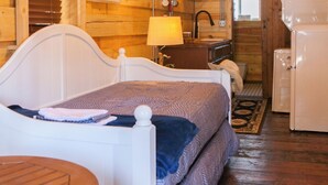 A wooden cabin interior with a bed, a small table, a kitchenette, and a bathroom. - Vintage Sheriff Tiny Cabin - HOT TUB! (Woodland Park)