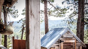 A rustic wooden outhouse with a metal roof, surrounded by tall pine trees and a mountainous backdrop. - Vintage Sheriff Tiny Cabin - HOT TUB! (Woodland Park)