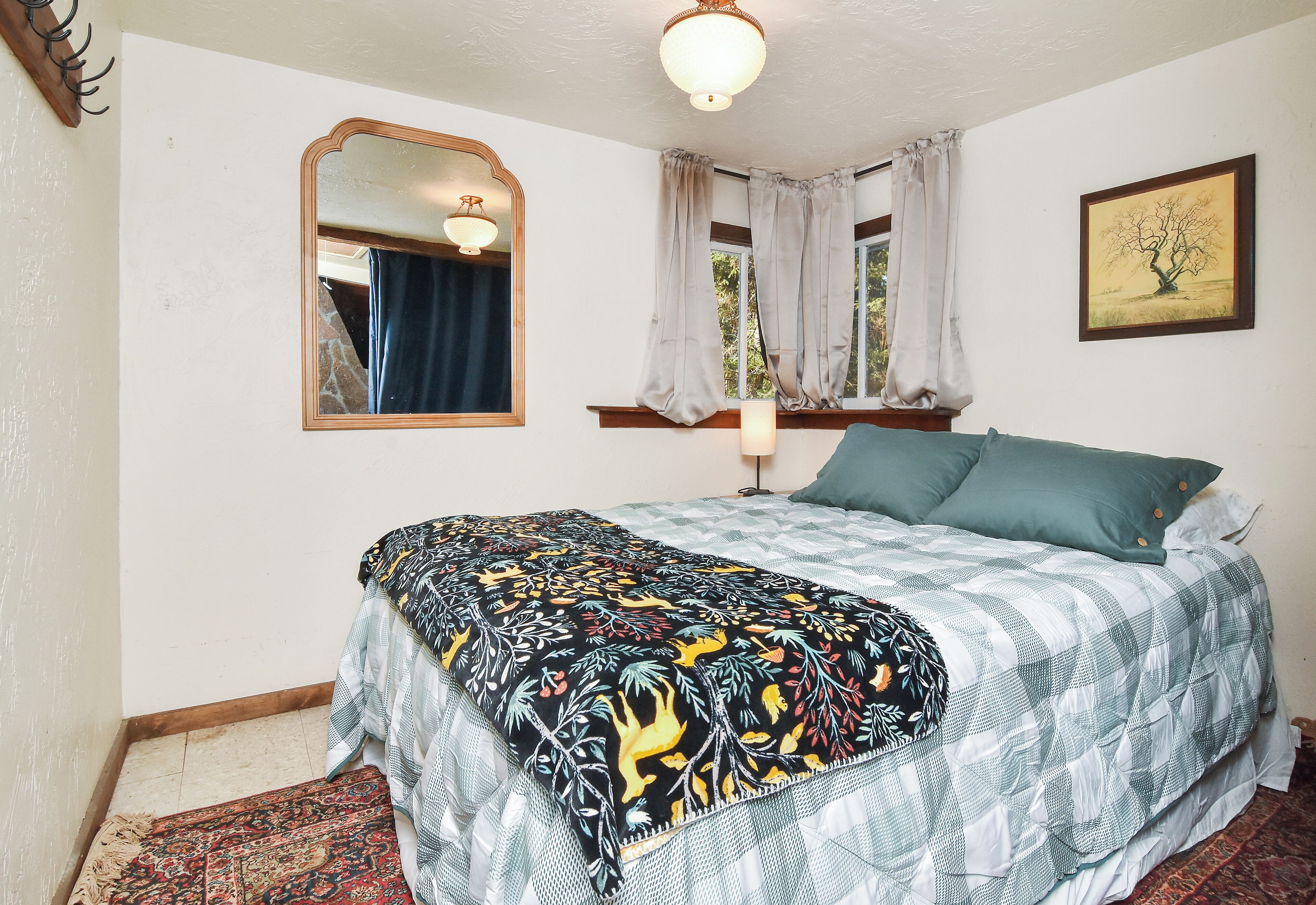 A bedroom with a bed, a window with curtains, a wall-mounted mirror, and a framed picture.