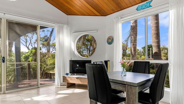 A dining area with a table and chairs, a round window with a tree view, and a sign indicating 'Beach' above a window. - Sails One - Inverloch (Inverloch)