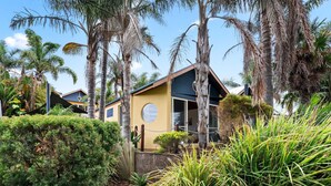 A yellow building with a circular window and a door, surrounded by palm trees and tropical vegetation. - Sails One - Inverloch (Inverloch)