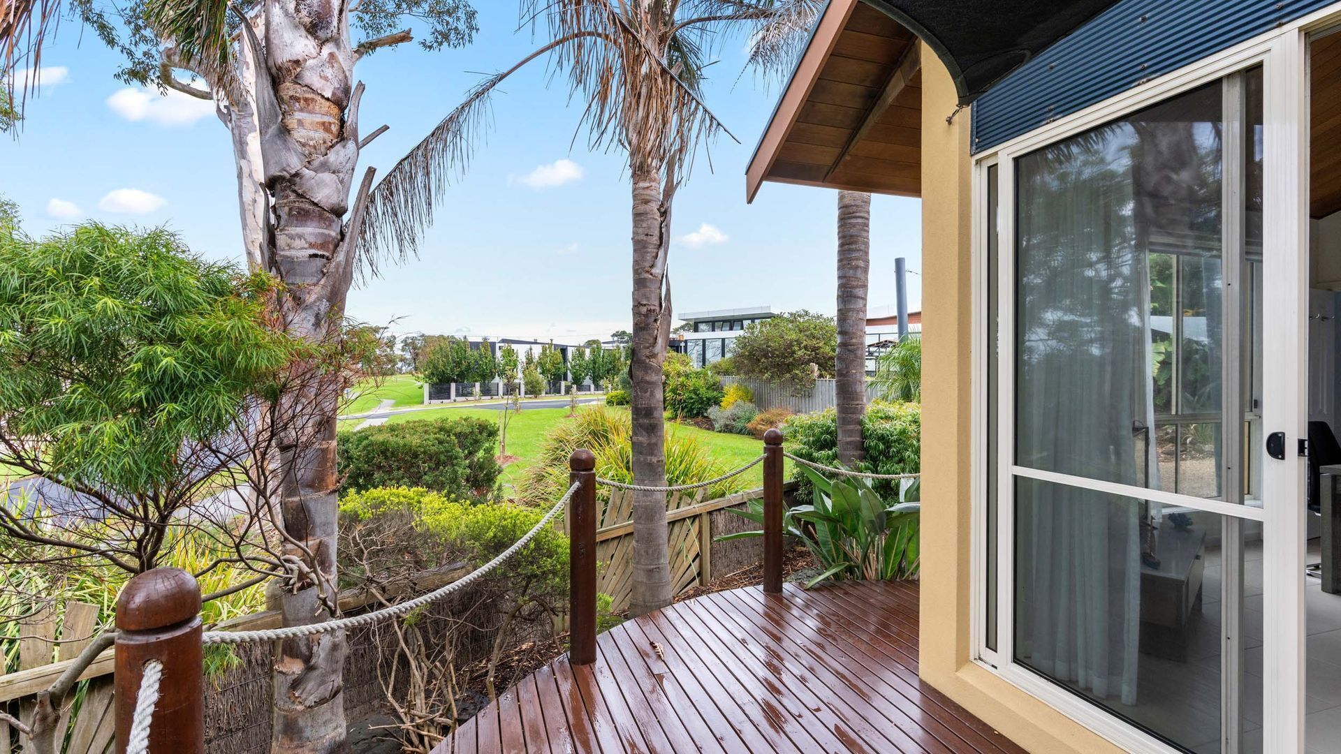 A wooden deck with a rope barrier, palm trees, and a view of a landscaped area with a building in the background.