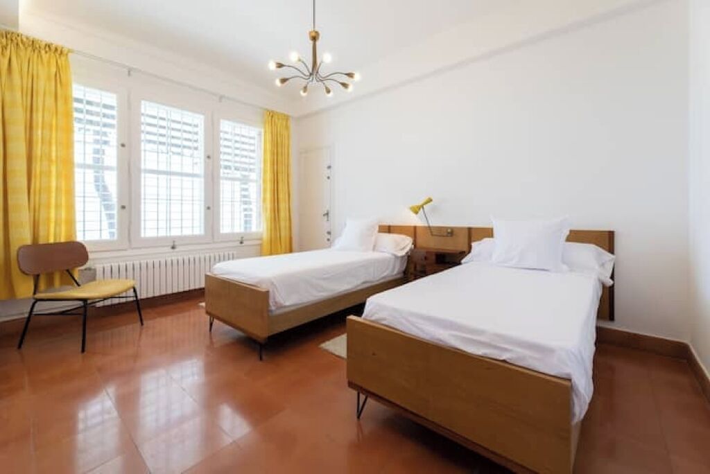 Une chambre d’hôtel avec deux lits simples, un plancher en bois, un radiateur, une chaise et une fenêtre avec des rideaux jaunes.