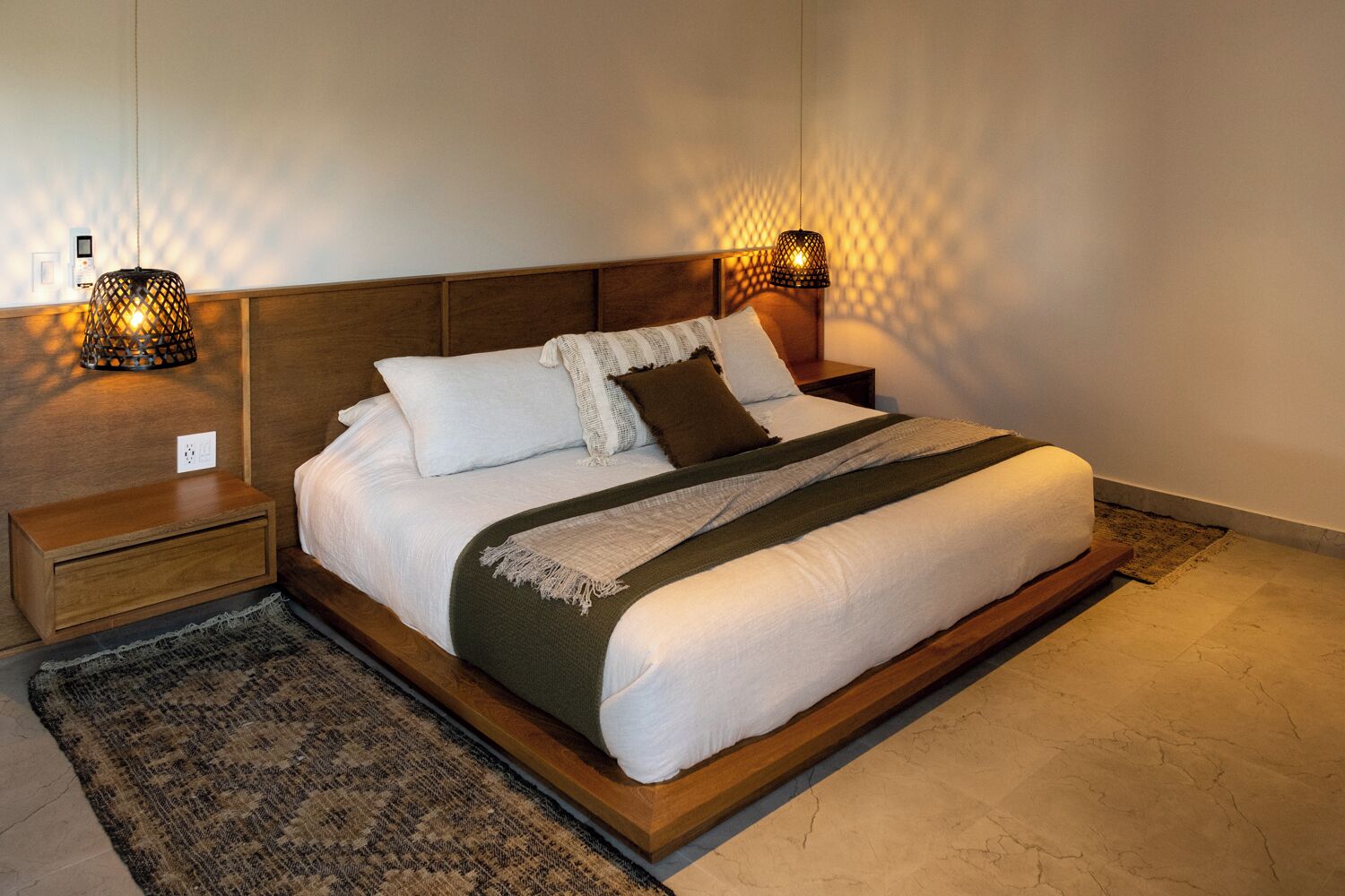Een net opgemaakt bed met een houten hoofdeinde, twee nachtkastjes met lampen, en een kamerbreed tapijt op de vloer.