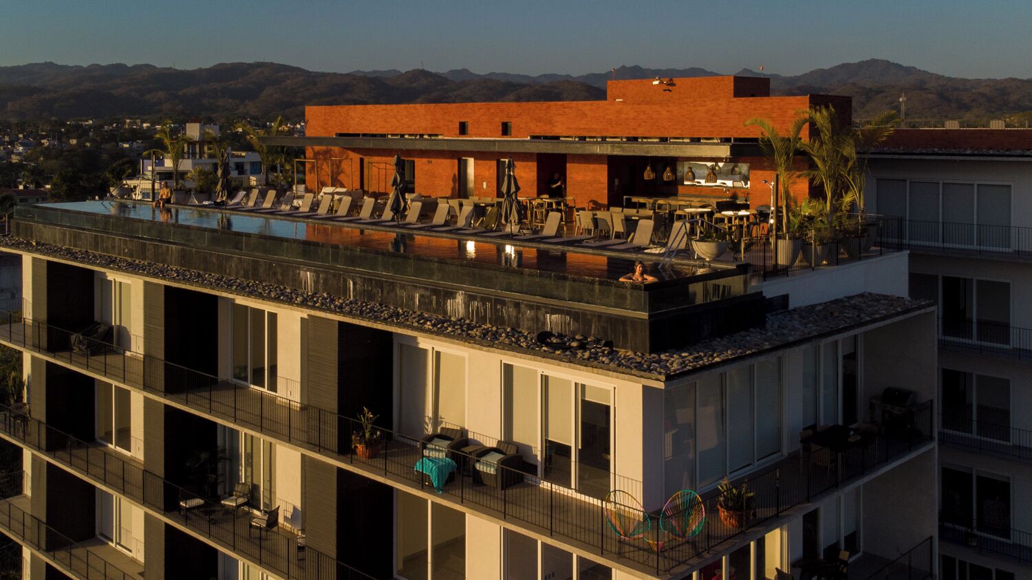 A modern building with a rooftop pool, multiple balconies, and a view of mountains in the background.
