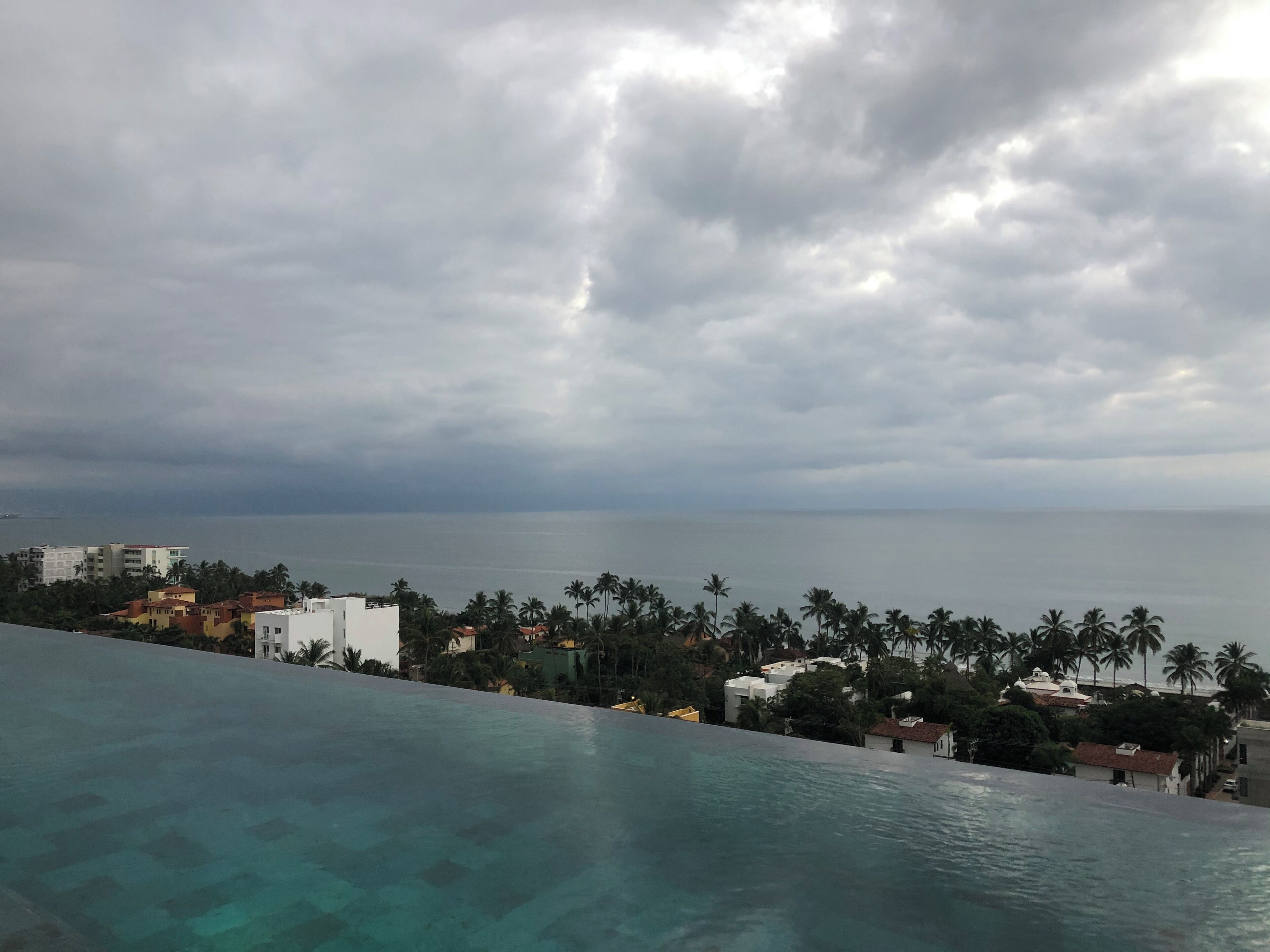A view of a swimming pool with a clear blue water surface, surrounded by buildings and palm trees, under a cloudy sky.