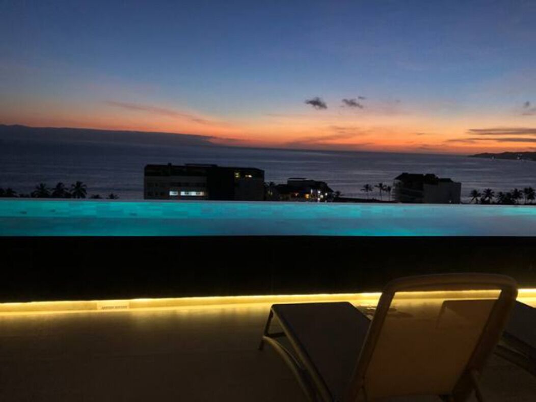 A poolside view at dusk with a sunset over the ocean, a lounge chair, and buildings in the background.