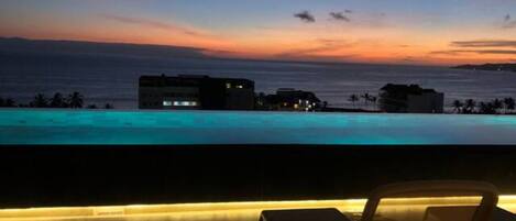 A poolside view at dusk with a sunset over the ocean, a lounge chair, and buildings in the background.