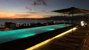 Vue nocturne d’un espace piscine avec des chaises longues et un parasol, donnant sur une étendue d’eau et sur le paysage urbain lointain.