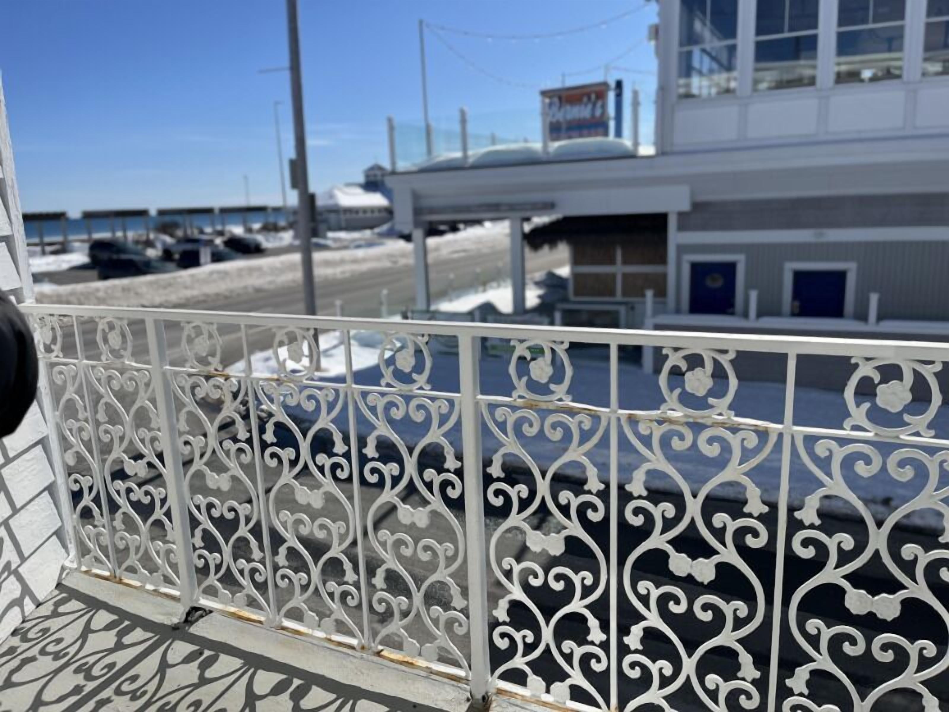 A metal balcony railing with intricate designs, overlooking a parking area and a building with a signboard.