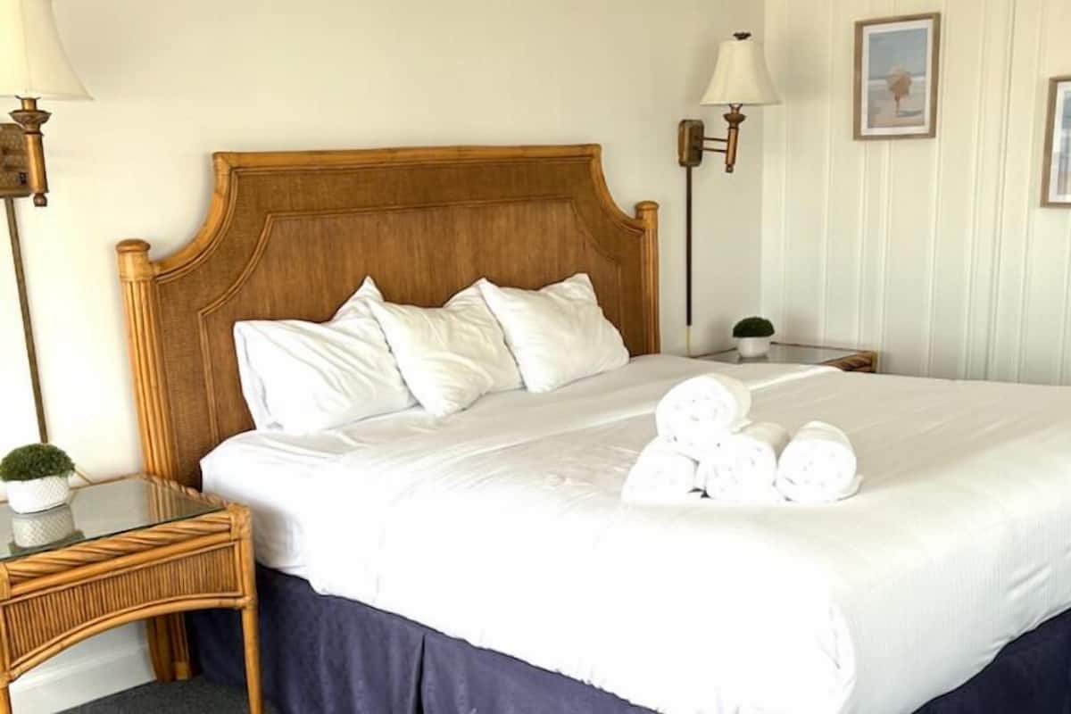 A neatly made bed with white linens and pillows, a wooden headboard, and a bedside table with a lamp and a small potted plant.