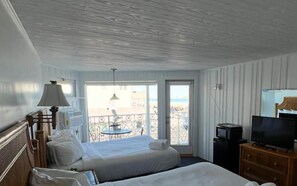 A hotel room with two beds, a TV, a small table, and a balcony with a view of the beach.
