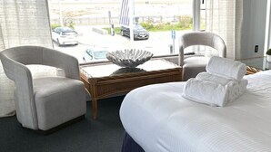 A hotel room with a large bed, two armchairs, a small coffee table, and a view of parked cars outside. - Harris Sea Ranch Beach Hotel (Hampton)