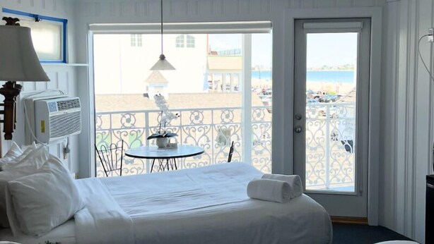 A hotel room with a bed, a table with chairs, a balcony with a railing, and a view of the beach. - Harris Sea Ranch Beach Hotel (Hampton)