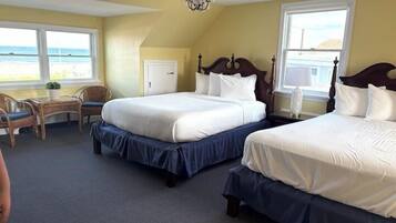 A hotel room with two beds, a desk, and a chair. There are large windows with a view of the beach.