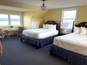 A hotel room with two beds, a desk, and a chair. There are large windows with a view of the beach.