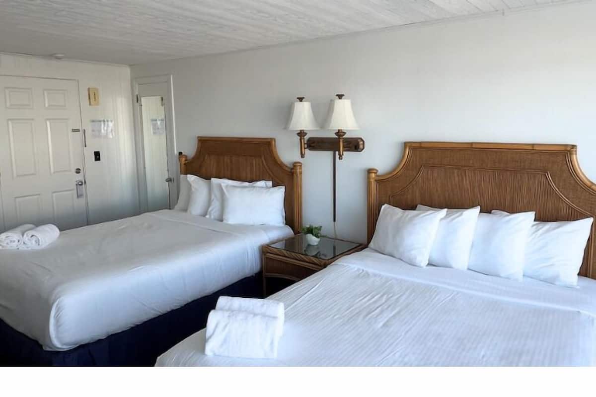 A hotel room with two beds, white linens, wooden headboards, a nightstand with lamps, and a door with a handle.