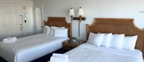 Habitación de hotel con dos camas, ropa de cama blanca, cabeceros de madera, una mesita de noche con lámparas y una puerta con picaporte.