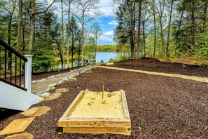 A wooden deck with a sandpit, surrounded by a forest and a lake.