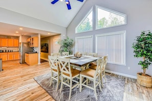 A dining area with a table and chairs, a kitchen in the background, and a large window with blinds.