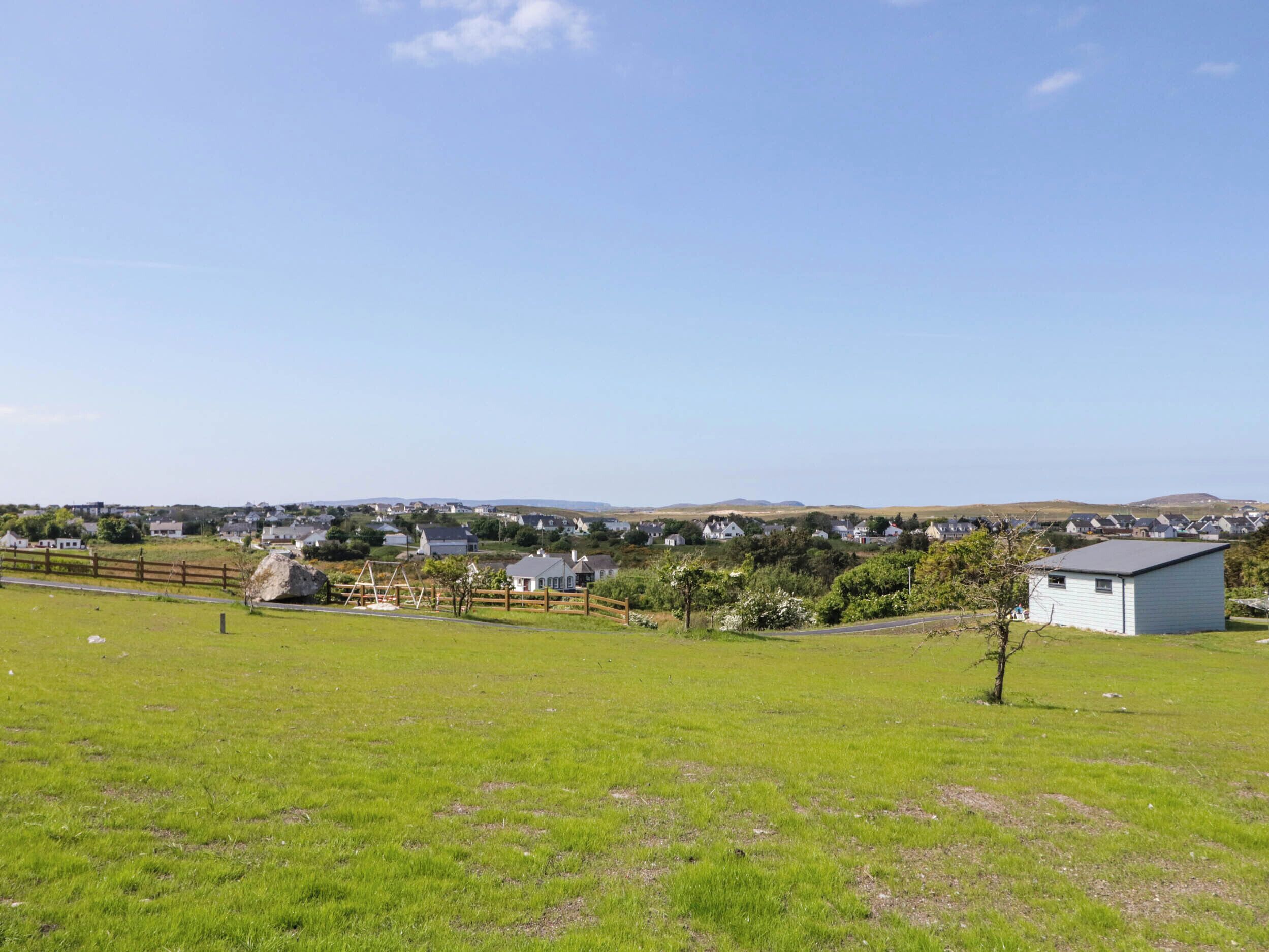 A grassy field with a wooden fence, a small shed, and houses in the background.