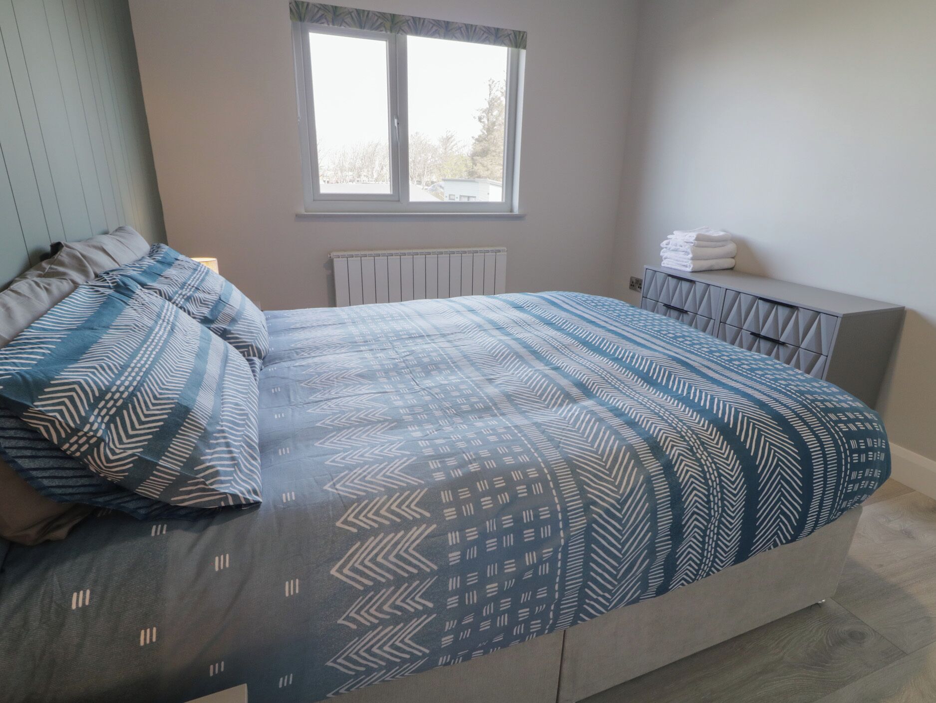 A bedroom with a bed, pillows, a window, a radiator, and a bedside table with towels.