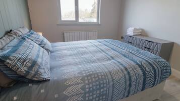 A bedroom with a bed, pillows, a window, a radiator, and a bedside table with towels.