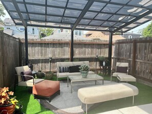 A covered patio with a wooden fence, outdoor furniture, and a glass table.