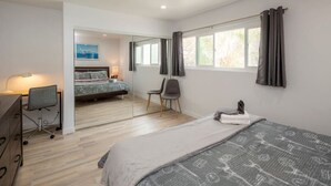 Een moderne slaapkamer met een groot bed, een bureau en een stoel. Er is een raam met gordijnen en uitzicht op groen.