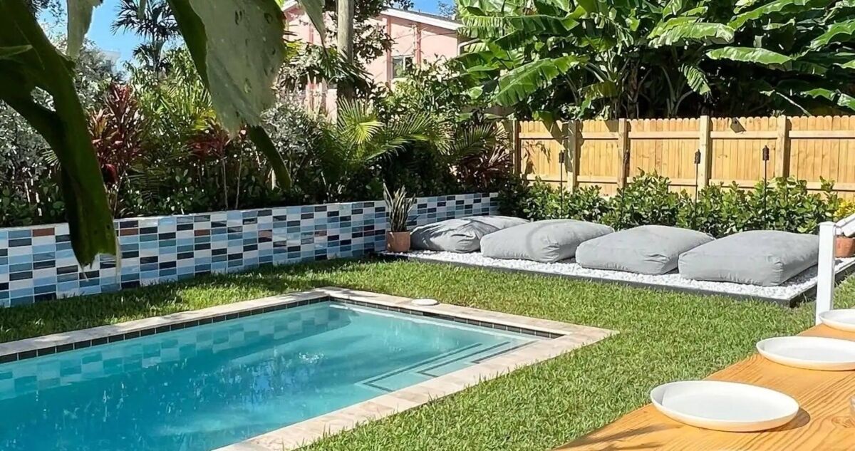 A swimming pool with a tiled border, surrounded by a grassy area and a wooden fence. There are several white cushions and a table with plates on it.