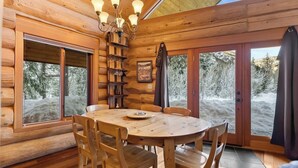 A wooden cabin interior with a round dining table, wooden chairs, a chandelier, and large windows offering a view of snow-covered trees. - River Front | Mountain Ski Cabin (Mazama)