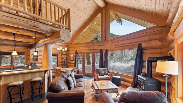 A log cabin interior with a wooden staircase, a dining area, a sofa, a fireplace, and large windows. - River Front | Mountain Ski Cabin (Mazama)