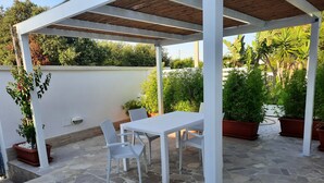 Un porche cubierto con una mesa y sillas blancas, rodeado de plantas en macetas y un suelo de piedra.