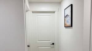 Un couloir étroit, avec une porte blanche au fond, un tableau encadré au mur et un plancher en bois.