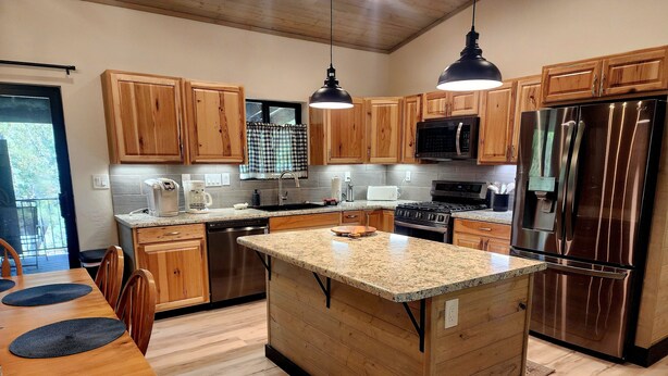 A modern kitchen with wooden cabinets, stainless steel appliances, a granite countertop island, and a dining area with wooden chairs. - BRAND NEW CUSTOM BUILT HOME WITH AC! FENCED FOR YOUR PUPS! (Pinetop-Lakeside)