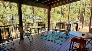 A covered outdoor deck with a table and chairs, surrounded by a wooden railing and overlooking a forested area. - BRAND NEW CUSTOM BUILT HOME WITH AC! FENCED FOR YOUR PUPS! (Pinetop-Lakeside)