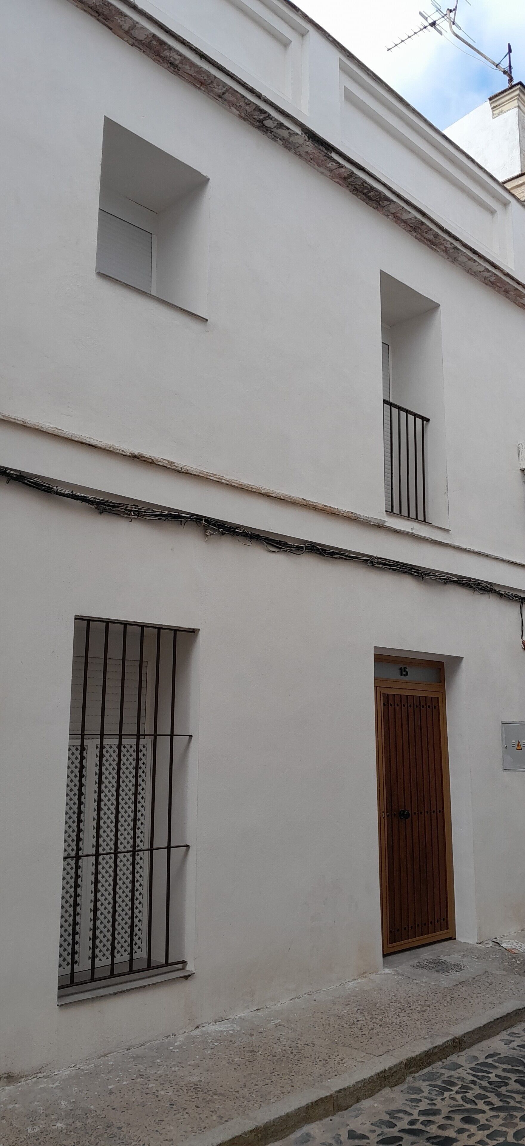 Un edificio blanco con puerta de madera marcada con el número 15, una ventana con barrotes y un pequeño balcón.