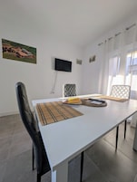 Een moderne eetruimte met een witte tafel, zwarte stoelen, een flatscreen-tv aan de muur en een ingelijst schilderij boven de tv.