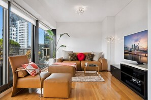 Un salon moderne avec un canapé, un fauteuil et un pouf, un téléviseur et une vue sur la ville.