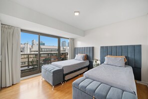 Habitación de hotel moderna con un ventanal grande, dos camas, una mesita pequeña y una mesita de noche.