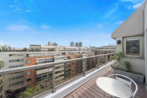Un balcón con una mesa y sillas de metal, vistas de edificios cercanos y un cielo despejado.