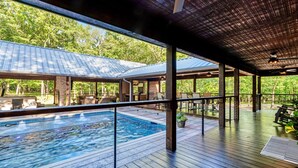 Zona de piscina exterior techada con terraza de madera, mobiliario de salón y vista a una casa al fondo.