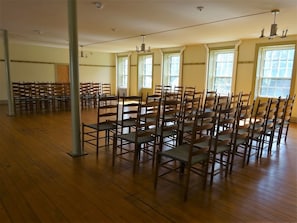 A spacious room with rows of wooden chairs and tables, large windows, and a high ceiling.