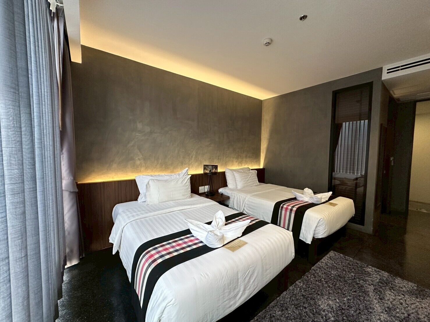 A hotel room with two beds, a dark floor, a large window with curtains, and a wall-mounted air conditioning unit.