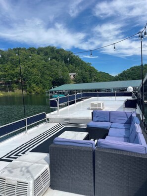 A boat deck with outdoor seating, a covered area, and a view of a forested area with a house in the background. - Houseboat Retreat on Norris Lake (Tazewell)