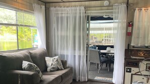 A cozy living area with a sofa, a patterned armchair, and a small table with a guitar. A window with curtains offers a view of the outdoors.